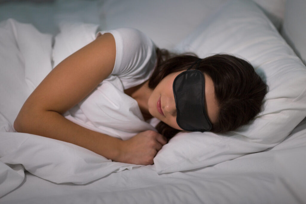 young woman sleeping with eye mask in bed at home › 스타일리쉬 플레이스 수면 안대를 착용하고 편안하게 잠든 여성. 목 통증 예방을 위한 적절한 수면 자세와 환경 조성의 중요성을 강조하는 장면.