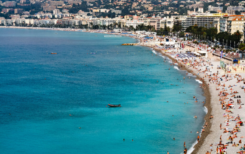 view on Azure coast in Nice › 스타일리쉬 플레이스 프랑스 니스의 해변 전경. 맑은 푸른 바다와 하얀 모래사장, 해변을 즐기는 사람들의 모습이 보입니다. 배경에는 도시의 건물들이 늘어서 있습니다.