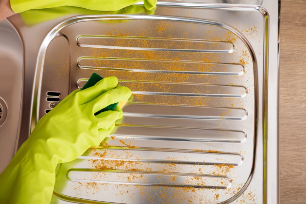 Woman Cleaning Untidy Stainless Steel Sink › 스타일리쉬 플레이스 고무장갑을 낀 손이 스테인리스 스틸 싱크대의 얼룩을 스펀지로 닦아내는 모습