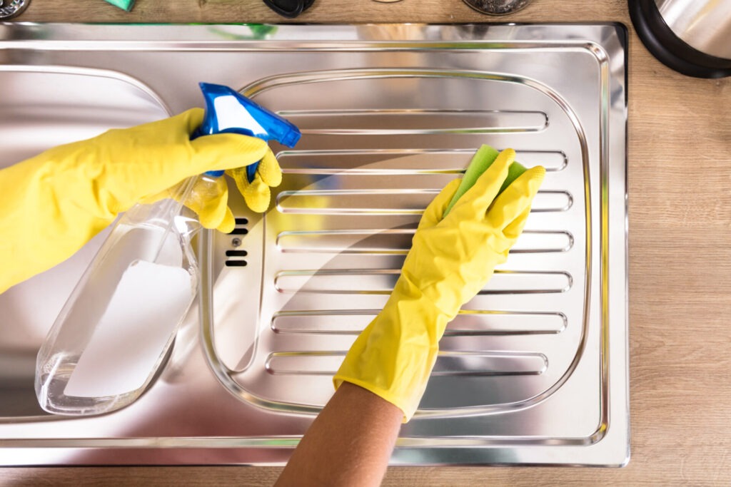 Persons Hand Cleaning Stainless Steel Sink › 스타일리쉬 플레이스 노란 고무장갑을 낀 손이 스프레이와 천을 사용해 스테인리스 스틸 싱크대를 닦는 모습