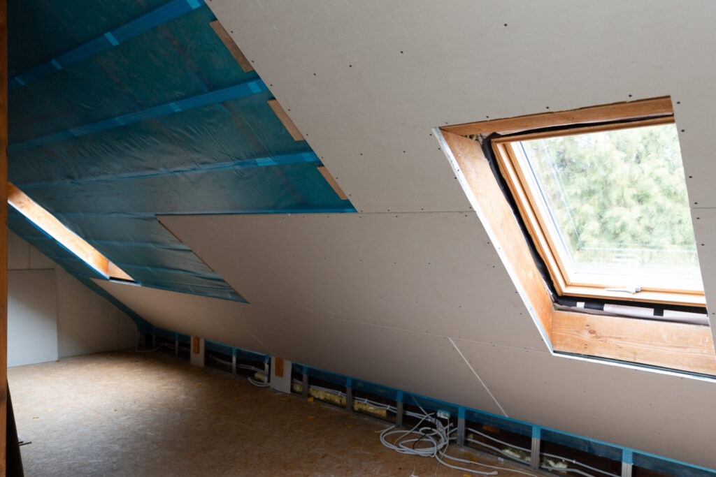 House attic insulation and renovation Drywall construction › 스타일리쉬 플레이스 지붕 아래 방의 내부 모습, 창문이 있는 경사진 천장. 적절한 단열과 환기가 곰팡이 발생을 방지하는 데 중요한 환경을 나타냅니다.