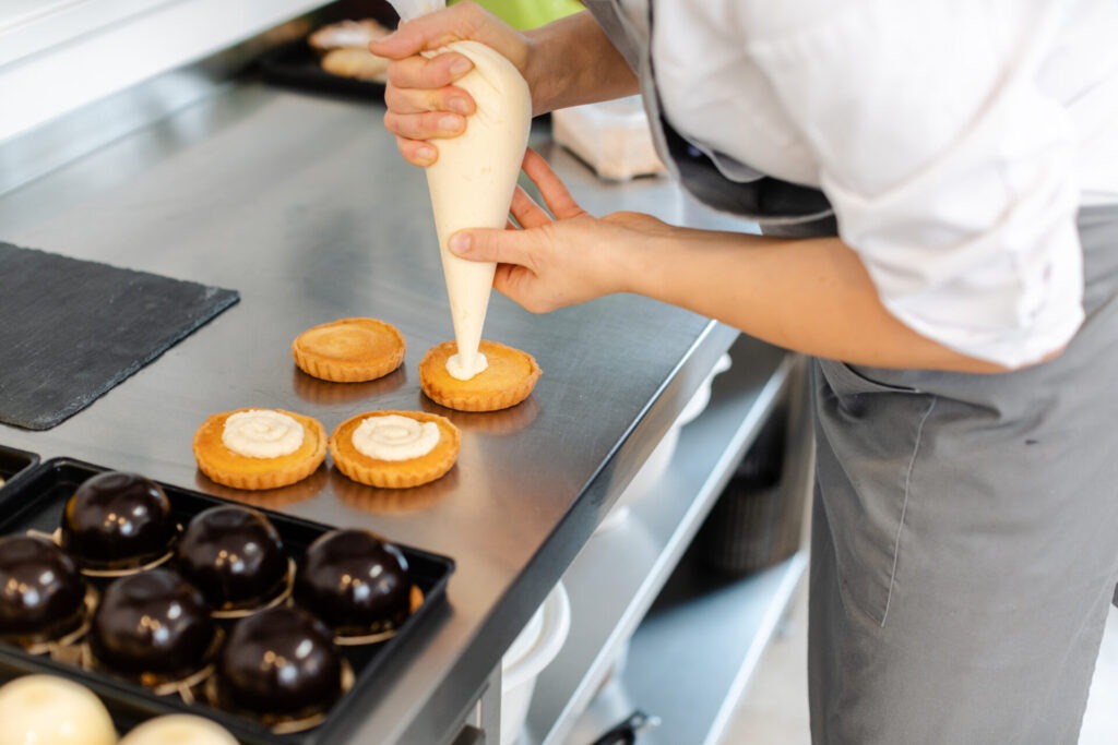 Pastry chef using icing bag to put creme on cake › 스타일리쉬 플레이스 크림을 짜는 제빵사의 손, 여러 개의 타르트가 놓인 작업대. 배경에는 다양한 디저트가 준비되어 있는 홈 베이킹 환경을 보여줍니다.
