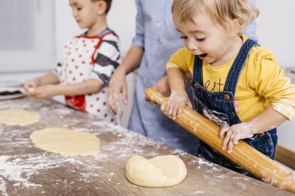 Excited toddler girl rolling out dough with wooden rolling pin › 스타일리쉬 플레이스 반죽을 밀고 있는 아이와 그 옆에서 지켜보는 또 다른 아이의 모습. 밀가루가 흩어져 있는 테이블에서 가족이 함께하는 따뜻한 베이킹 장면.