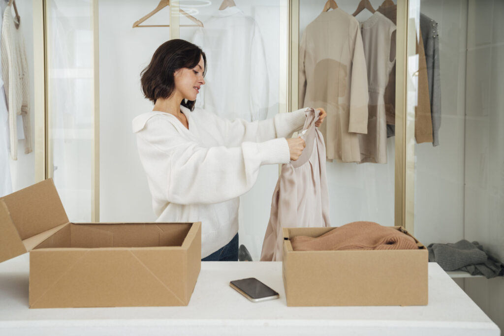 Young woman recycling clothes with box on table at home › 스타일리쉬 플레이스 가을 옷 정리와 관리를 위해 옷을 정리하는 장면을 담은 이미지로, 계절별 의류 보관 및 정리 팁에 적합합니다.