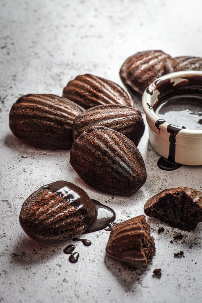 Schokoladen-Madeleines mit Schokoladendip › 스타일리쉬 플레이스 다양한 초콜릿 마들렌이 놓여 있는 테이블, 일부는 초콜릿 소스에 담겨 있으며, 한 개는 조각난 모습. 부드러운 질감과 풍부한 맛을 강조하는 이미지.