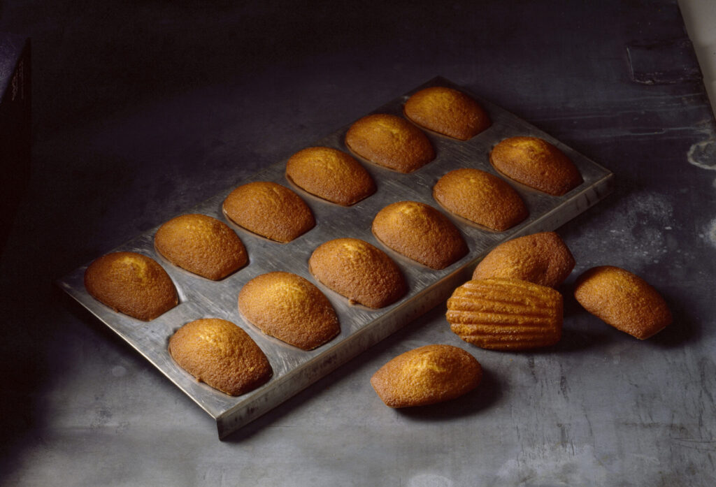 removing madeleine sponge cakes from tin › 스타일리쉬 플레이스 금색으로 구워진 마들렌이 팬에 정렬되어 있는 모습. 일부는 팬에서 떨어져 있으며, 부드러운 질감과 고소한 색상이 돋보이는 이미지.