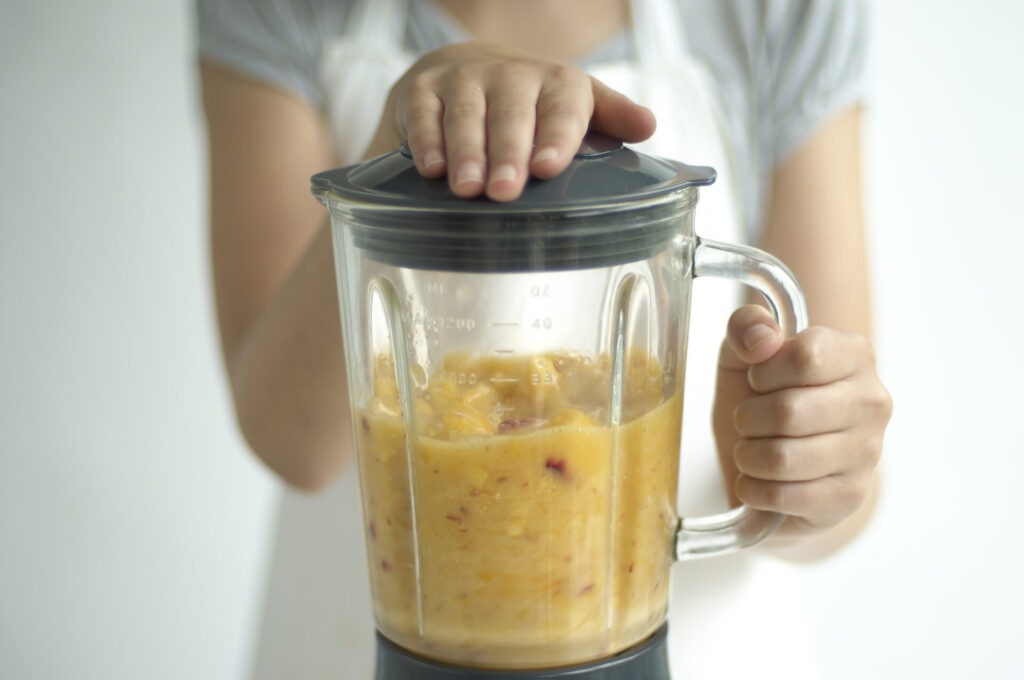 Child holding blender containing peach puree › 스타일리쉬 플레이스 과일 스무디를 만들고 있는 손이 블렌더의 뚜껑을 올리고 있는 모습. 블렌더 내부에는 갈아진 과일이 들어 있습니다.
