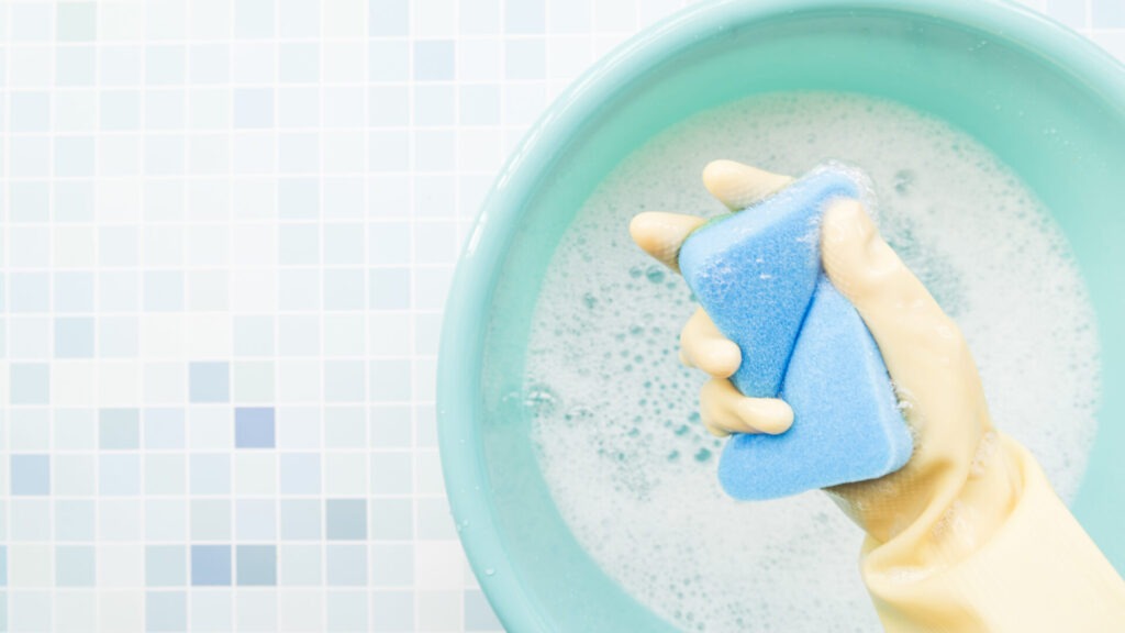 hand holding sponge › 스타일리쉬 플레이스 거품이 있는 물이 담긴 그릇에서 파란 스펀지를 쥐고 있는 손. 청소 과정을 나타내며, 효과적인 벽 청소 방법을 암시하는 이미지.