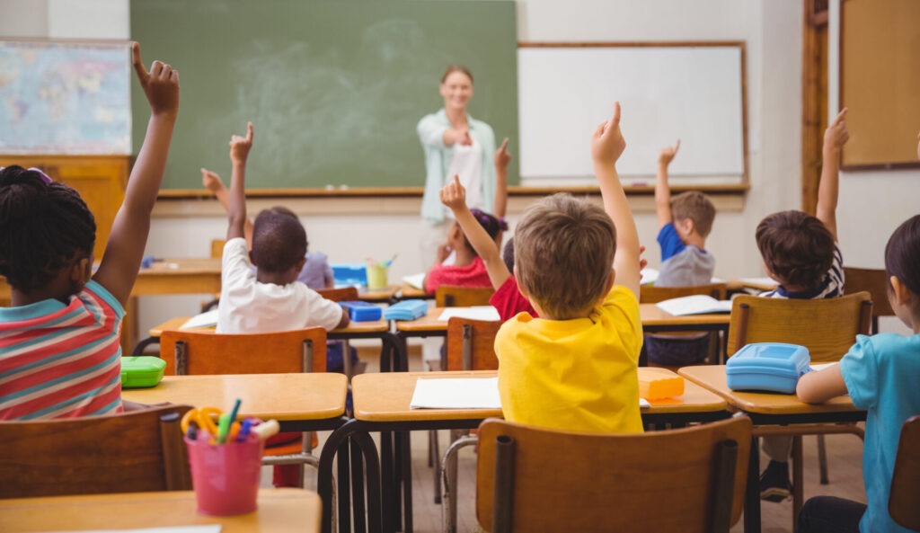 Pupils raising their hands during at the elementary school › 스타일리쉬 플레이스 교실에서 교사가 학생에게 질문을 하고, 학생들이 손을 들어 답변하고 싶어 하는 모습. 다양한 인종의 학생들이 자유롭게 의견을 표현하며 독립성을 기르는 장면.