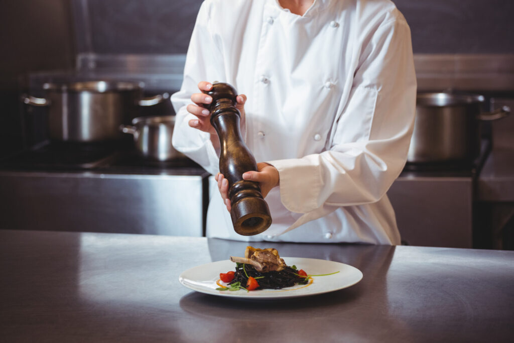 Chef sprinkling pepper on dish in a commercial kitchen › 스타일리쉬 플레이스 흰색 조리복을 입은 셰프가 후추통을 들고 요리 위에 후추를 뿌리고 있는 모습. 배경에는 조리 중인 냄비들이 보입니다.
