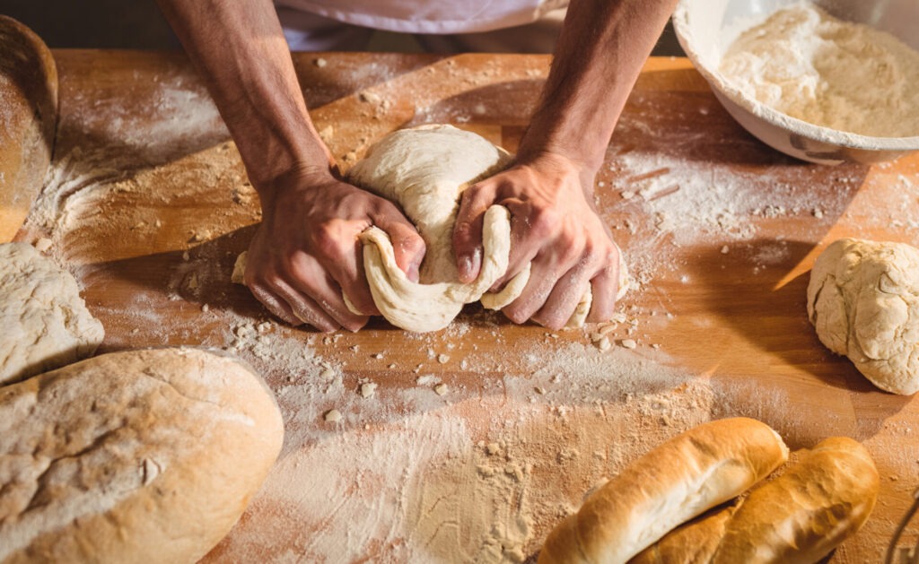 Hands of baker kneading a dough in bakery shop › 스타일리쉬 플레이스 밀가루가 뿌려진 나무 테이블 위에서 반죽을 손으로 치대고 있는 모습. 주변에는 다양한 형태의 빵이 놓여 있으며, 홈 베이킹의 과정을 보여줍니다.