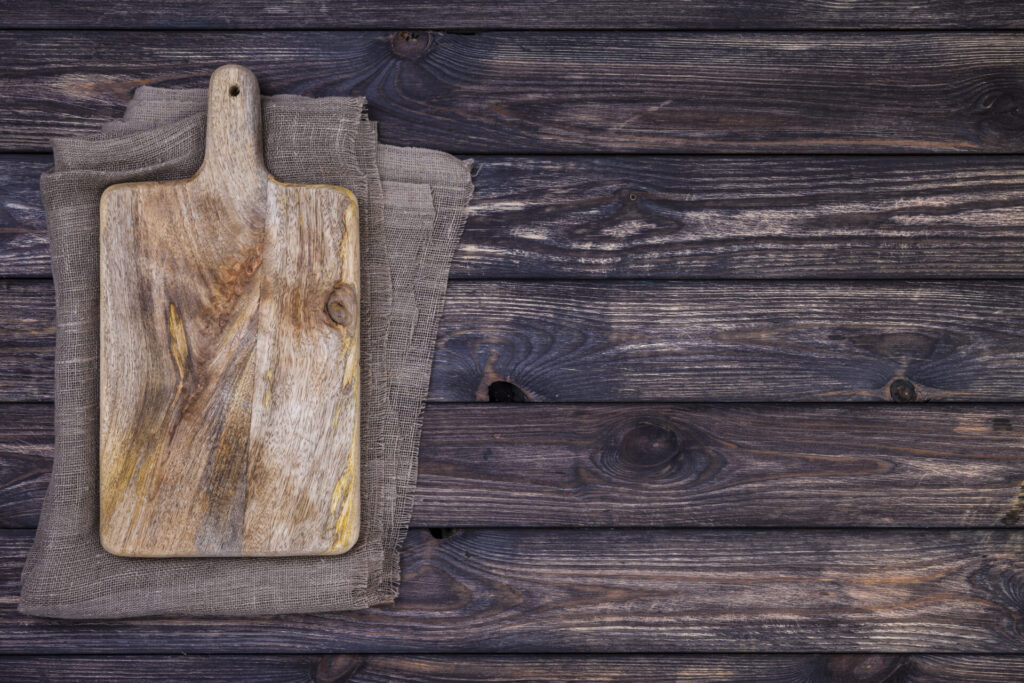 Old cutting board on dark wooden table Top view Copy space › 스타일리쉬 플레이스 어두운 나무 배경 위에 놓인 나무 도마가 보인다. 도마는 자연스러운 질감과 색상을 지니고 있으며, 그 아래에는 베이지색의 천이 깔려 있다.