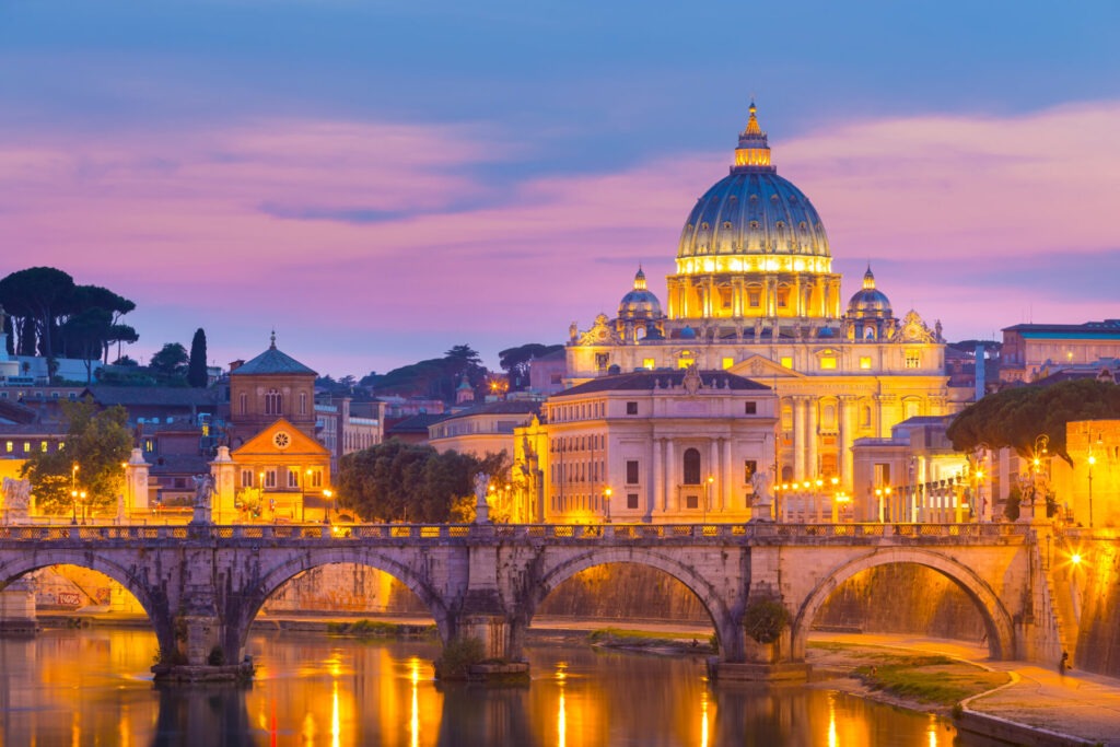 View at St Peters cathedral in Rome Italy › 스타일리쉬 플레이스 저녁 노을이 지는 로마의 전경. 성 베드로 대성당의 둥근 지붕과 밝은 조명, 티베르 강 위의 다리가 보입니다. 하늘은 보라색과 주황색으로 물들어 있습니다.