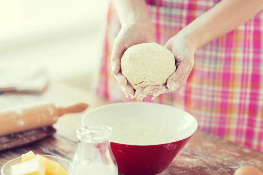 close up of female hands kneading dough at home › 스타일리쉬 플레이스 빨간 그릇에서 반죽을 손으로 들고 있는 모습. 주변에는 밀가루와 버터, 계란이 놓여 있으며, 홈 베이킹의 따뜻한 분위기를 나타냅니다.