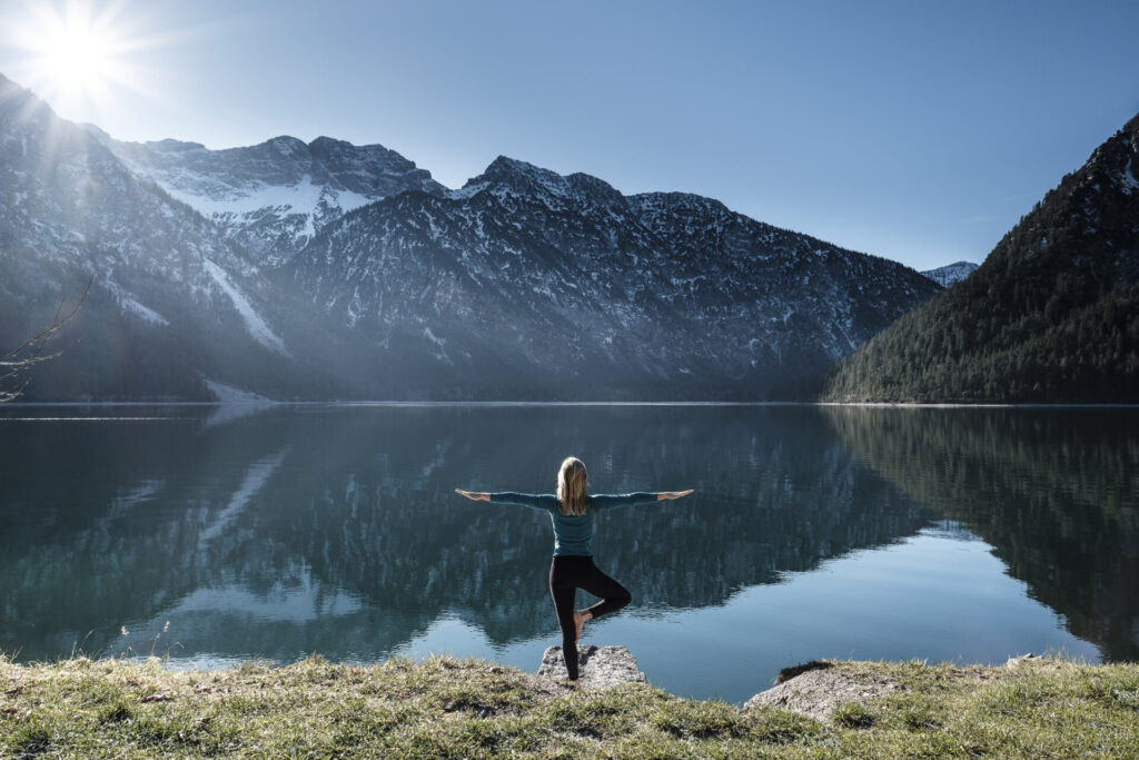 Woman practicing yoga at Lake Plansee Ammergau Alps Reutte Tyrol Austria › 스타일리쉬 플레이스 산과 호수를 배경으로 요가를 하는 여성, 자연 속에서 마음의 평화를 찾는 모습