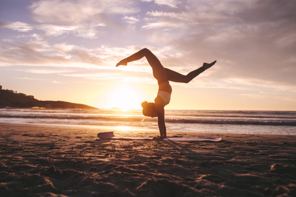 Woman doing yoga pose handstand on beach › 스타일리쉬 플레이스 해변에서 요가를 하는 사람, 일몰 배경 속에서 명상과 요가로 마음의 평화를 찾는 모습