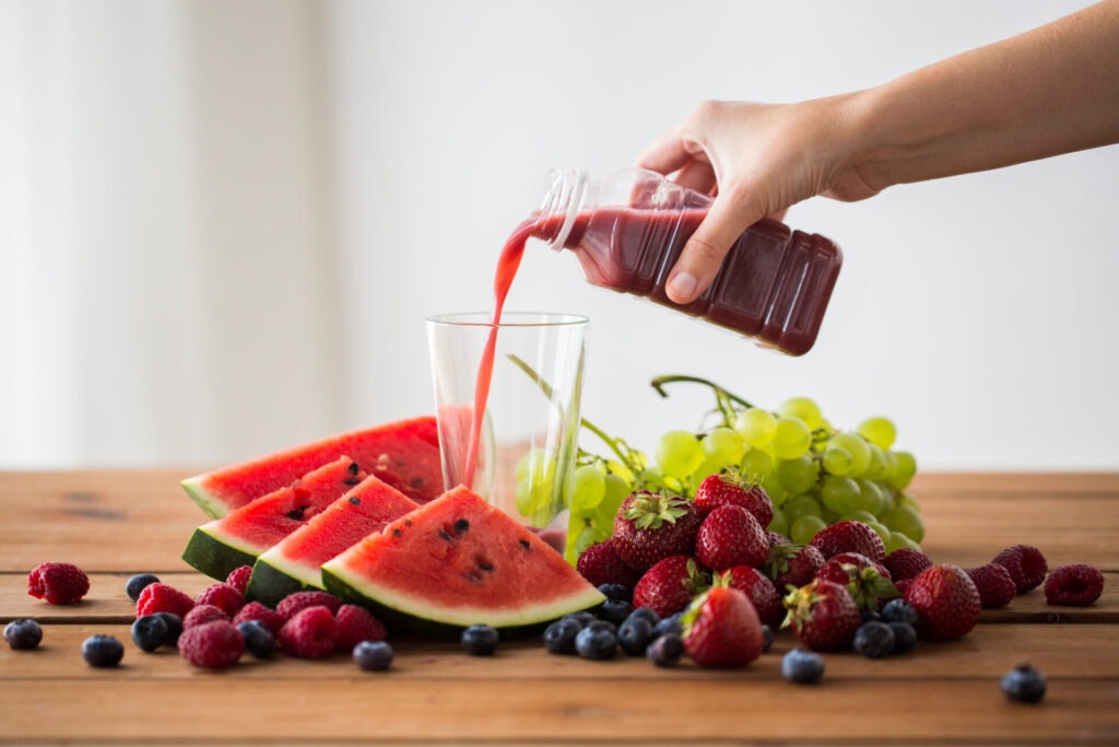hand pouring fruit juice from bottle to glass › 스타일리쉬 플레이스 유리컵에 빨간 과일 주스를 붓고 있는 모습, 주변에 수박, 딸기, 포도 등 다양한 과일이 놓여 있음