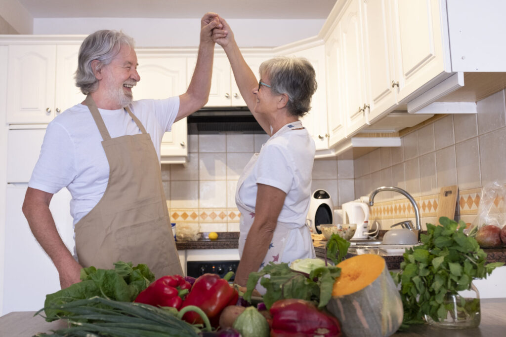 Senior couple holding hand while dancing in kitchen › 스타일리쉬 플레이스 부엌에서 신선한 채소를 앞에 두고 손을 맞잡은 시니어 부부, 비건 요리를 준비하며 기쁨을 나누는 모습