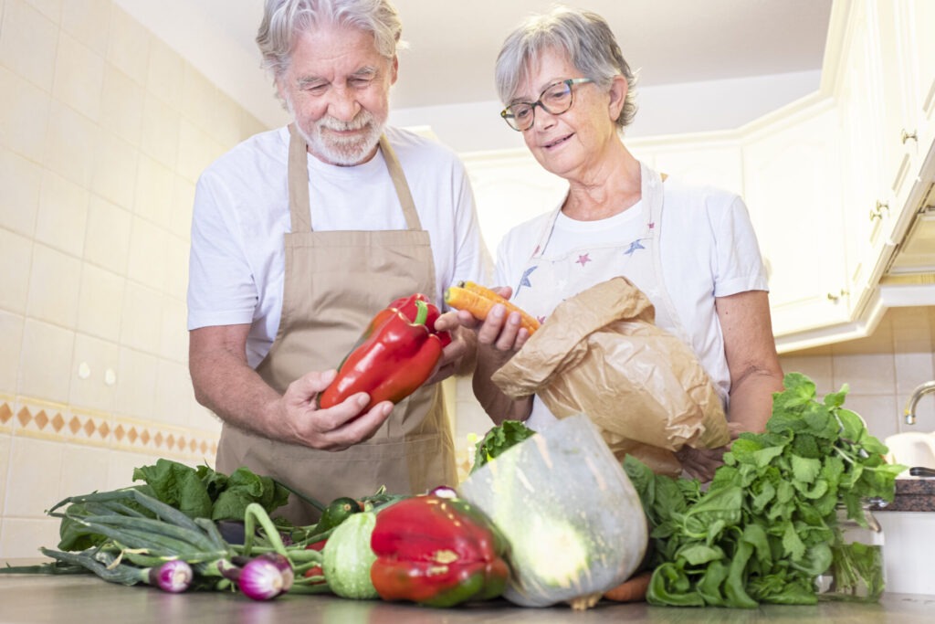 Senior couple removing vegetables from paper bag in kitchen › 스타일리쉬 플레이스 주방에서 빨간 파프리카와 당근 등 다양한 신선 채소를 고르는 시니어 부부, 비건 식생활 준비 모습