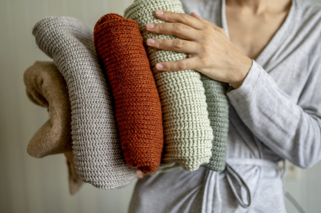 Woman holding clean folded sweaters at home › 스타일리쉬 플레이스 겨울옷 정리 예시 사진 - 여러 색상의 니트를 손에 들고 정리하는 모습
