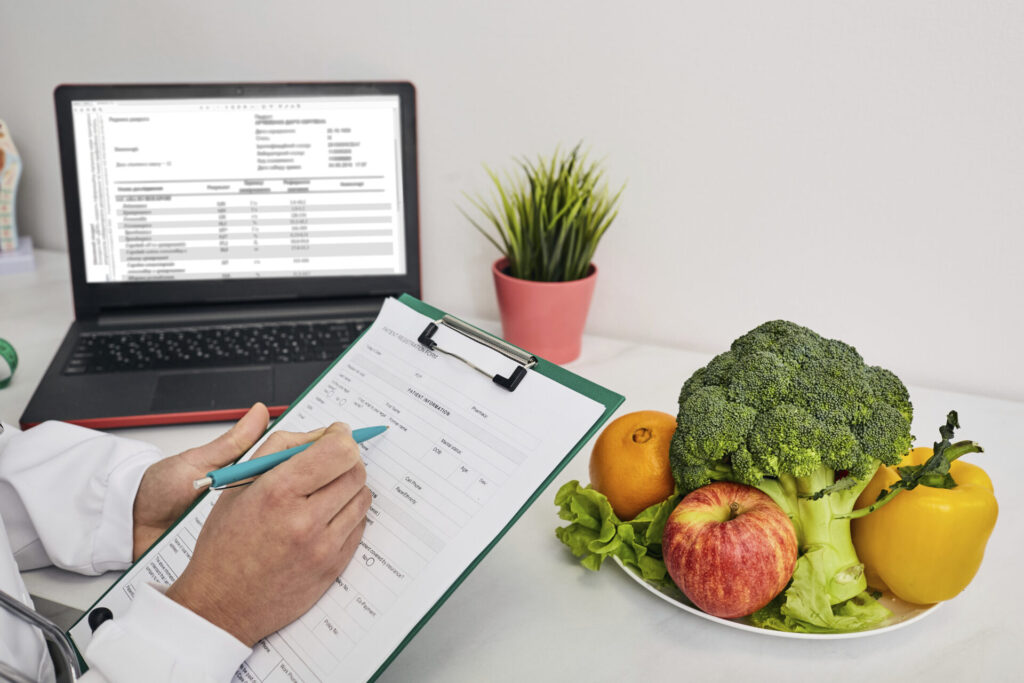 Nutritionist consultation › 스타일리쉬 플레이스 면역력 증진에 좋은 브로콜리와 과일이 담긴 접시를 앞에 두고, 식단 기록지를 작성 중인 의료 전문가 사진