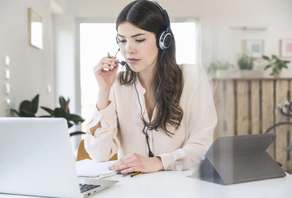 Young woman sitting at table at home wearing a headset › 스타일리쉬 플레이스 헤드셋을 착용한 여성이 노트북 앞에서 화상 회의하며 재택근무 중 메모를 작성하는 모습