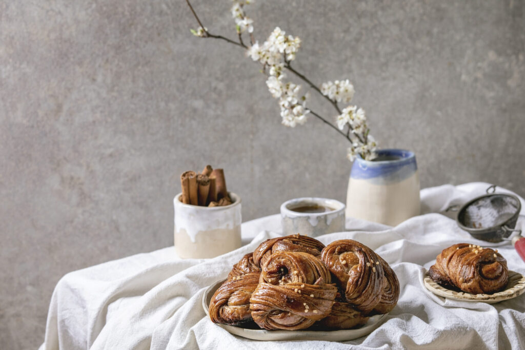 Swedish cinnamon sweet buns › 스타일리쉬 플레이스 화이트 테이블보 위에 놓인 시나몬 트위스트 빵 한 접시와 뒤편에 놓인 꽃 가지 및 차