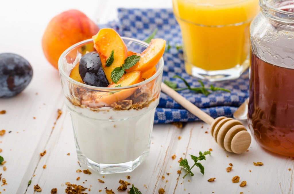 Greek yogurt with fresh fruit › 스타일리쉬 플레이스 복숭아와 자두 슬라이스, 그래놀라, 요거트가 담긴 유리컵 디저트와 꿀, 주스가 함께 놓인 테이블 세팅 이미지