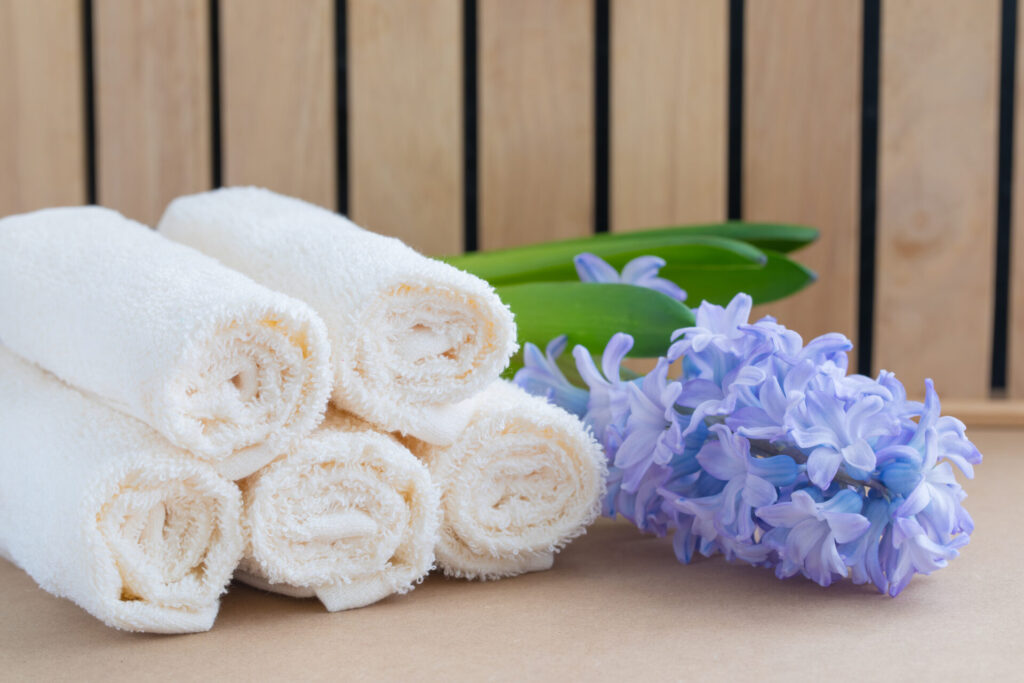 Beige bath towels on wooden background and a fresh violet hyacinth Spa concept › 스타일리쉬 플레이스 말끔하게 말린 흰색 타월과 보랏빛 꽃이 함께 놓인 이미지, 친환경 세탁 후의 청결한 느낌